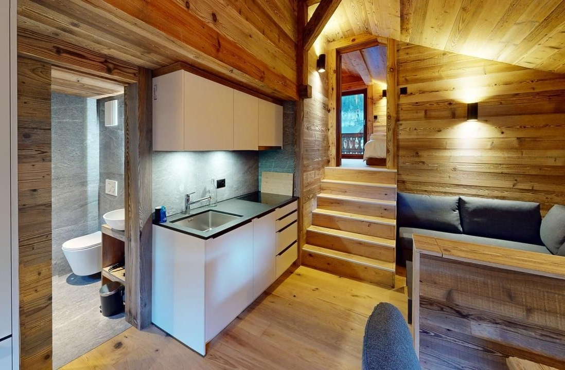 A rustic cabin interior featuring a kitchenette with white cabinets, a bathroom entrance, stairs, and a grey sofa.