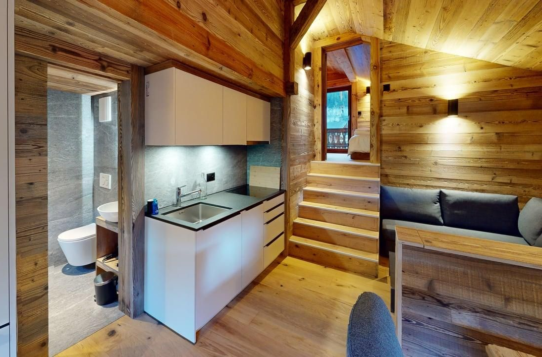 A wooden cabin interior featuring a modern kitchenette, a small staircase, a couch, and a view into a bathroom.