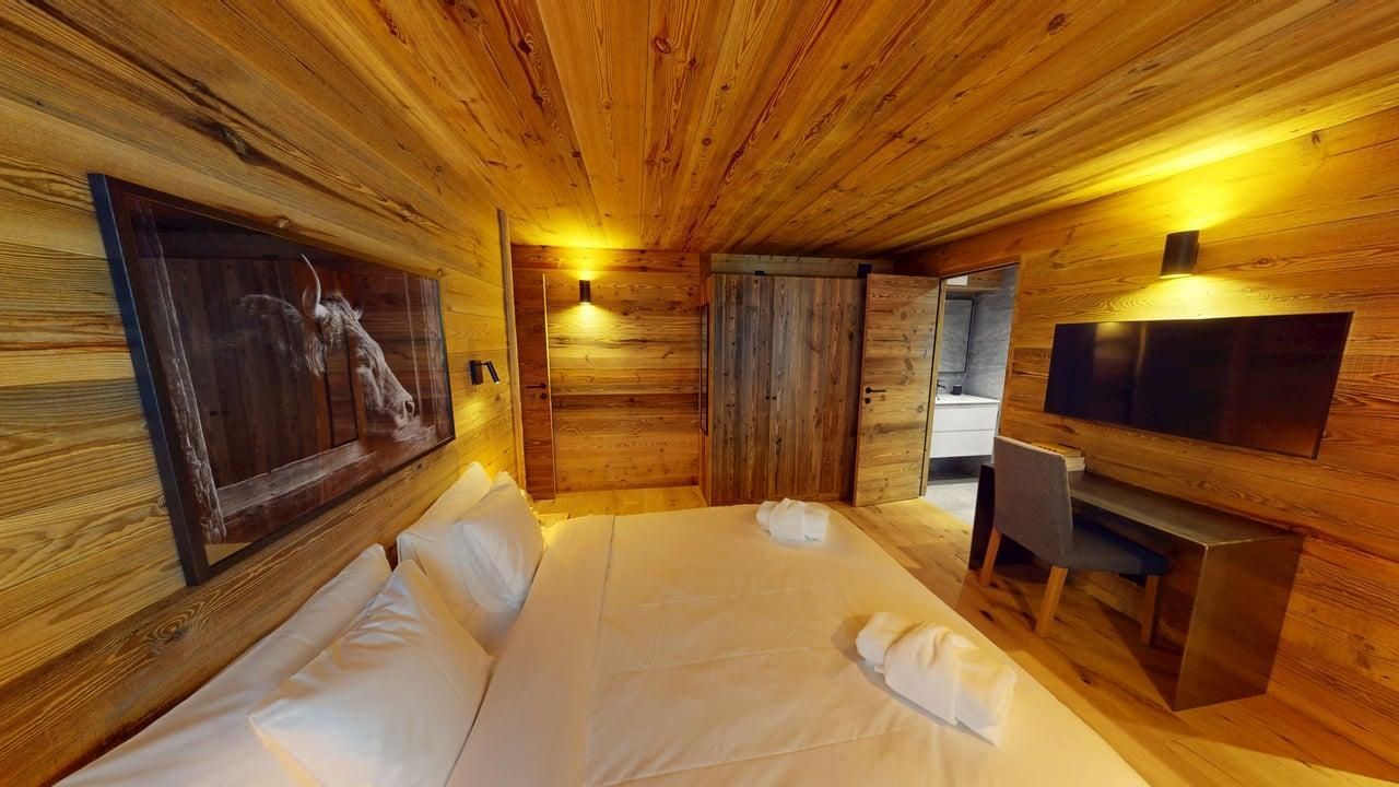 A rustic bedroom with wooden walls and ceiling, a neatly made bed, a desk with a chair, and a framed wall artwork.