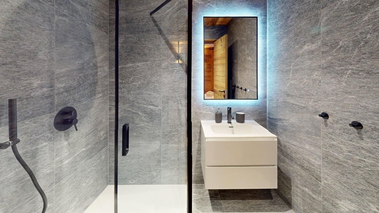 Salle de bains moderne avec murs en pierre grise, douche à l'italienne en verre, meuble-lavabo suspendu blanc et miroir rétroéclairé.