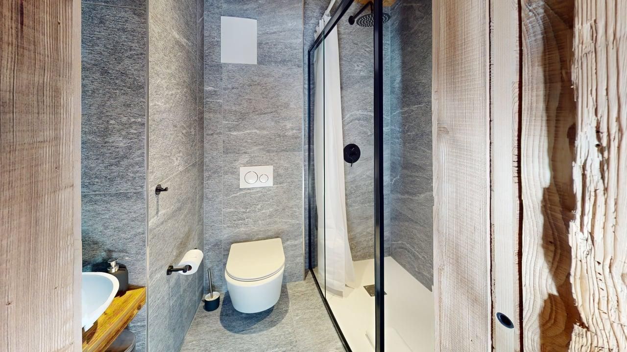 A modern bathroom featuring gray stone walls, a white toilet, a walk-in shower with a glass door, and wooden accents.