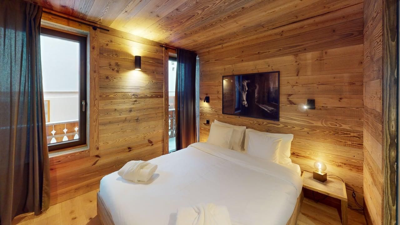 Une chambre confortable aux murs et au plafond en bois, avec un lit aux draps blancs et des fenêtres donnant sur un balcon.