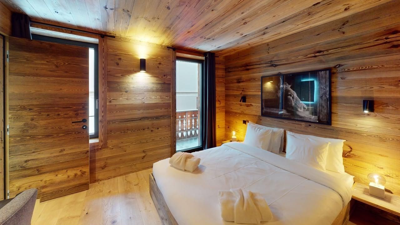 Une chambre rustique avec des murs lambrissés, un grand lit aux draps blancs et un balcon visible par une fenêtre.