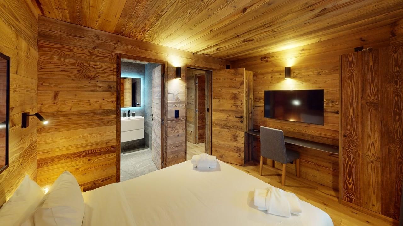 A rustic, wood-paneled hotel room with a neatly made bed, wall-mounted TV, desk, chair, and a view into a modern bathroom.