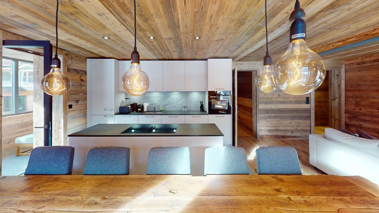 Cuisine moderne avec une table en bois, des chaises bleues, un îlot central et trois grands luminaires suspendus dans une pièce de style rustique.