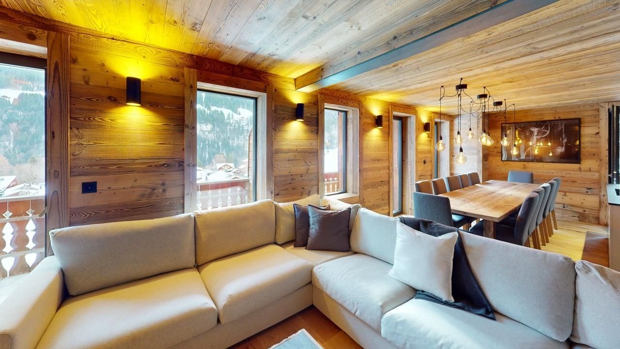 A modern living and dining room with a large, beige sectional sofa, rustic wooden walls, and a long dining table.
