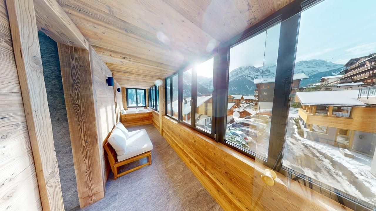 Un balcon étroit, lambrissé de bois, avec un siège blanc donnant sur les montagnes enneigées et les chalets du village.