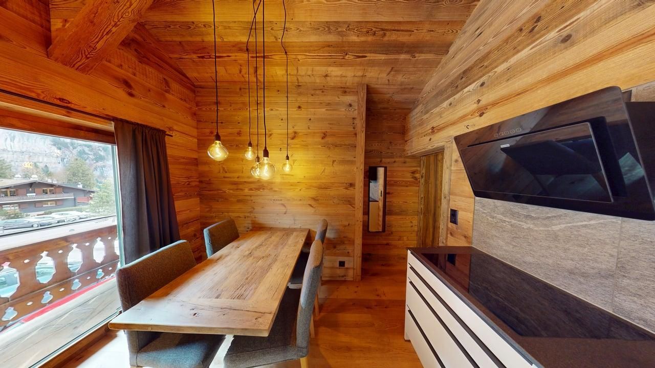 Une salle à manger rustique en bois avec une longue table, des chaises grises, des luminaires suspendus et un meuble de cuisine moderne.