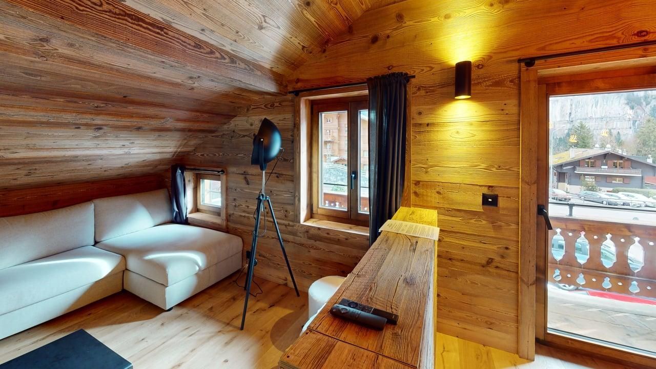 Un salon mansardé confortable est doté de murs lambrissés, d'un canapé d'angle blanc, d'une lampe trépied et d'une vue sur un balcon.
