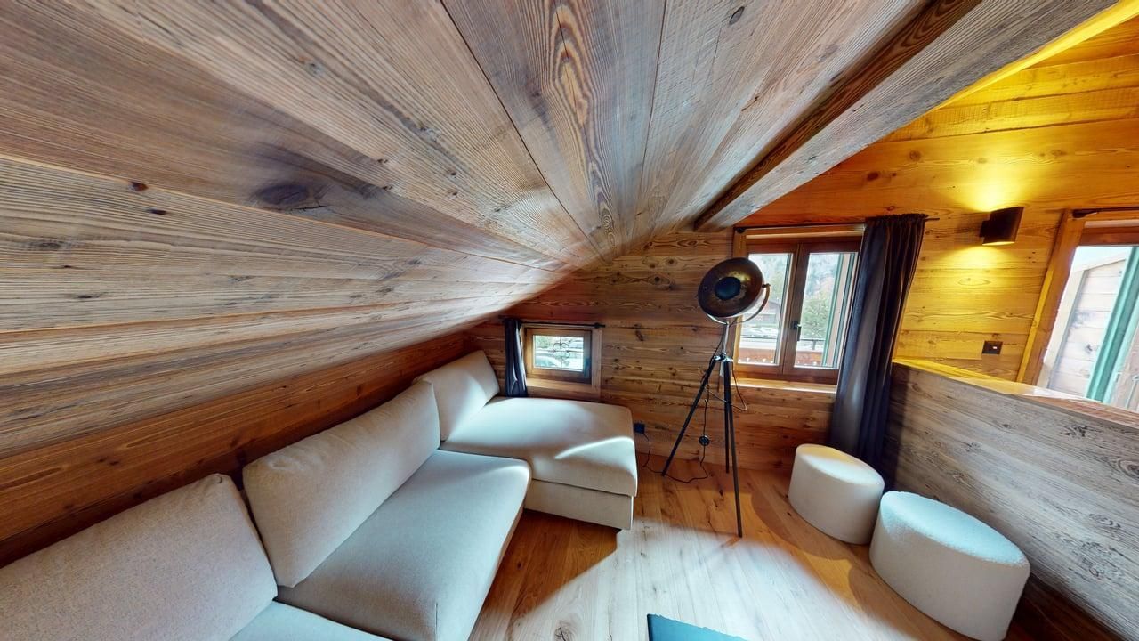 Un coin salon mansardé confortable avec un canapé d'angle beige, deux petits tabourets, des murs en bois, un plafond mansardé et un lampadaire.