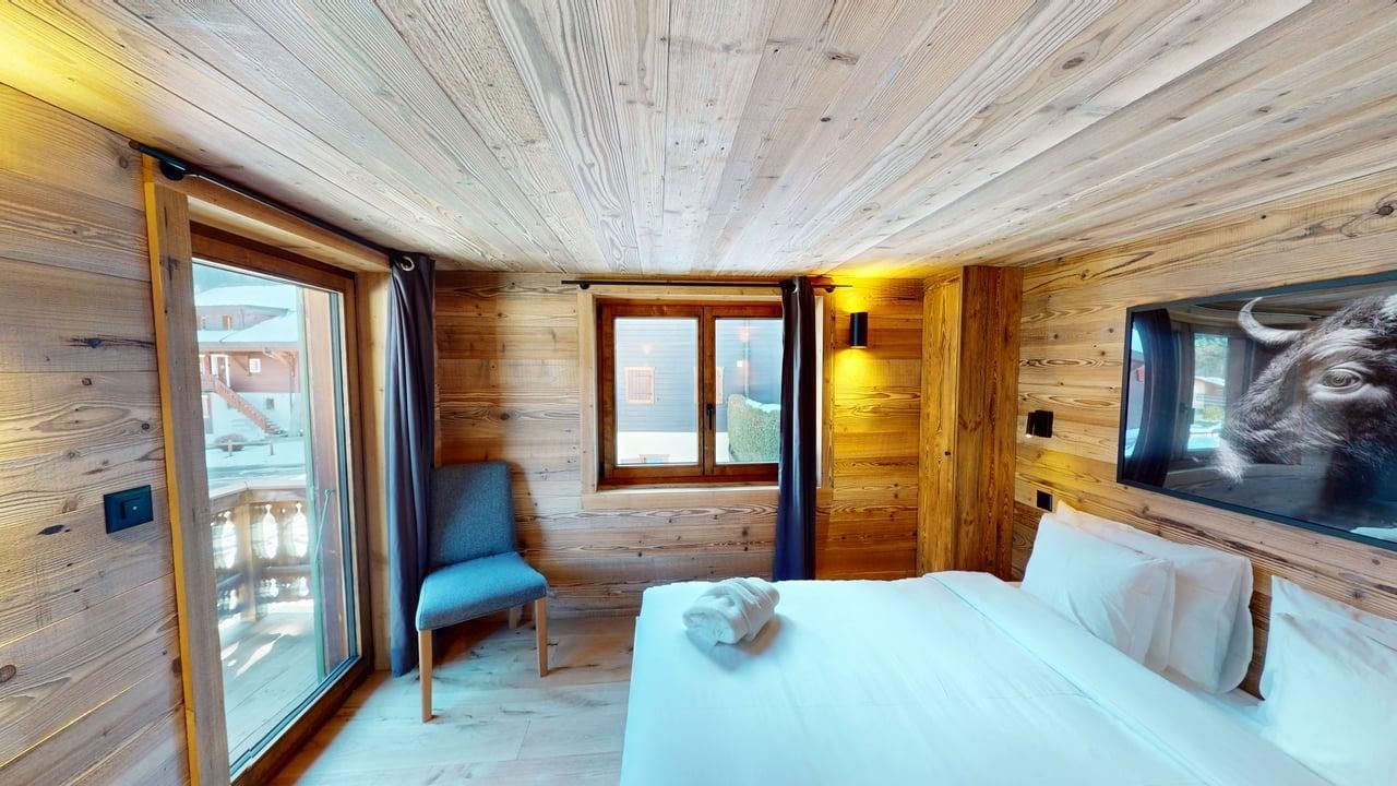 Une chambre confortable avec des murs et un plafond lambrissés, un lit blanc, un fauteuil bleu près d'une fenêtre et une télévision fixée au mur.