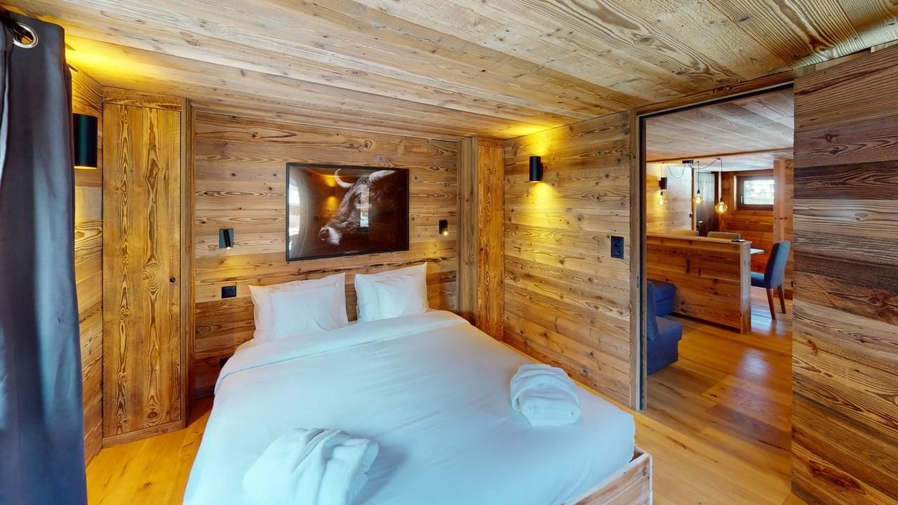 Une chambre rustique avec des murs lambrissés, un lit aux draps blancs et une porte ouverte donnant sur un autre espace de vie.
