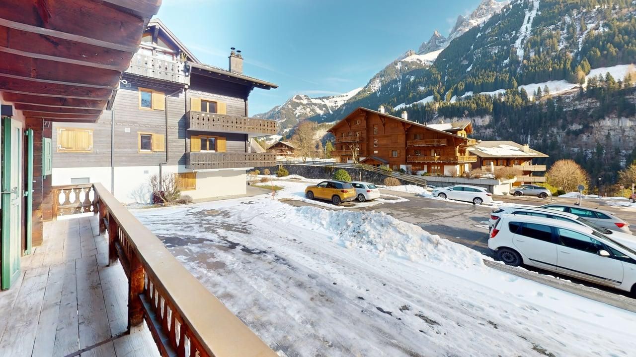Un parking enneigé entouré de chalets de montagne traditionnels en bois, avec en arrière-plan des sommets enneigés.