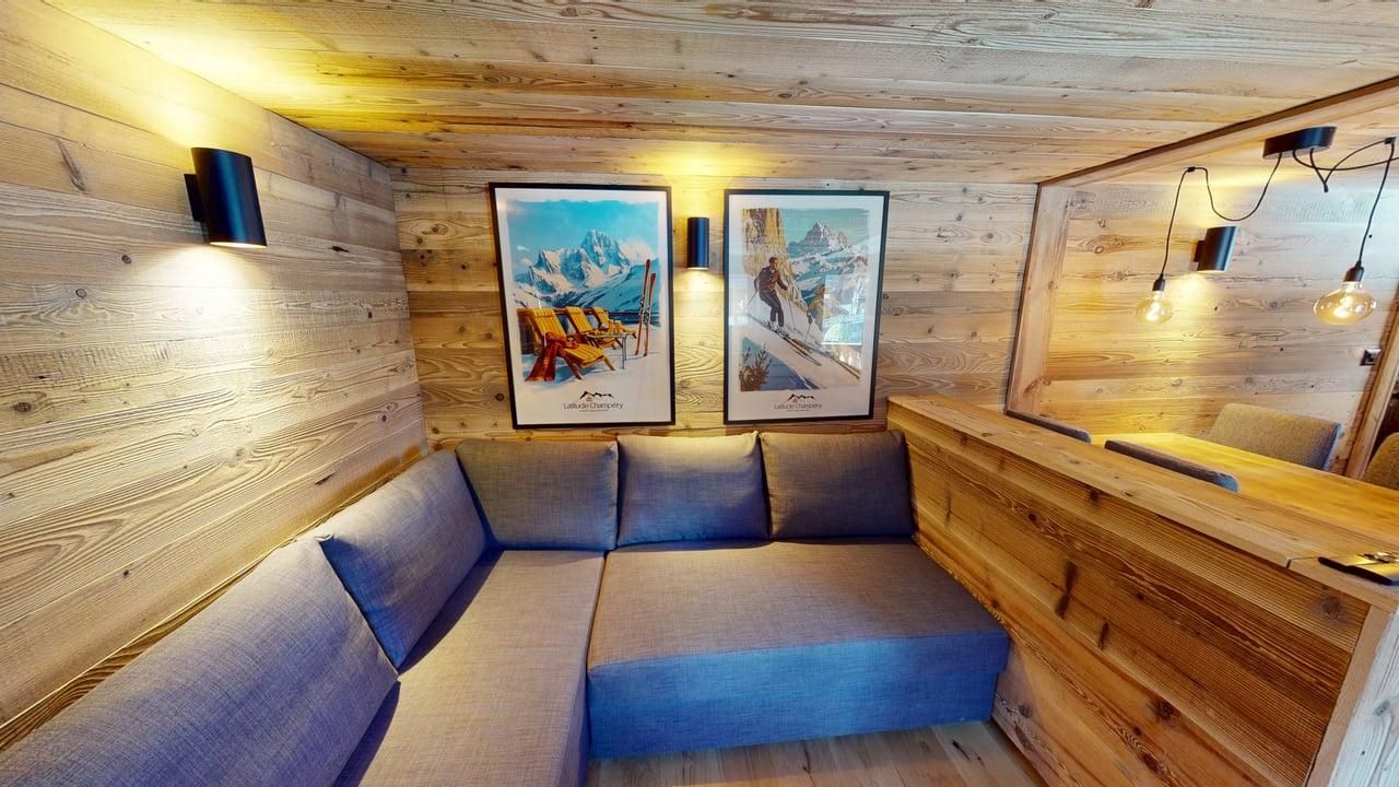 Une pièce confortable aux boiseries chaleureuses comprend un canapé d'angle bleu, deux reproductions encadrées de paysages de montagne et un éclairage tamisé.
