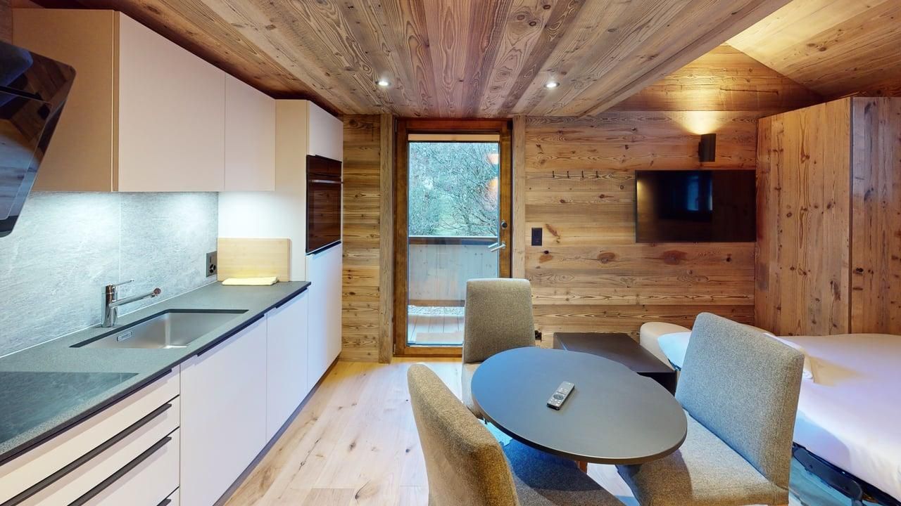 Une chambre d'hôtel moderne, lambrissée, avec une petite kitchenette, une table à manger avec deux chaises et une télévision murale.