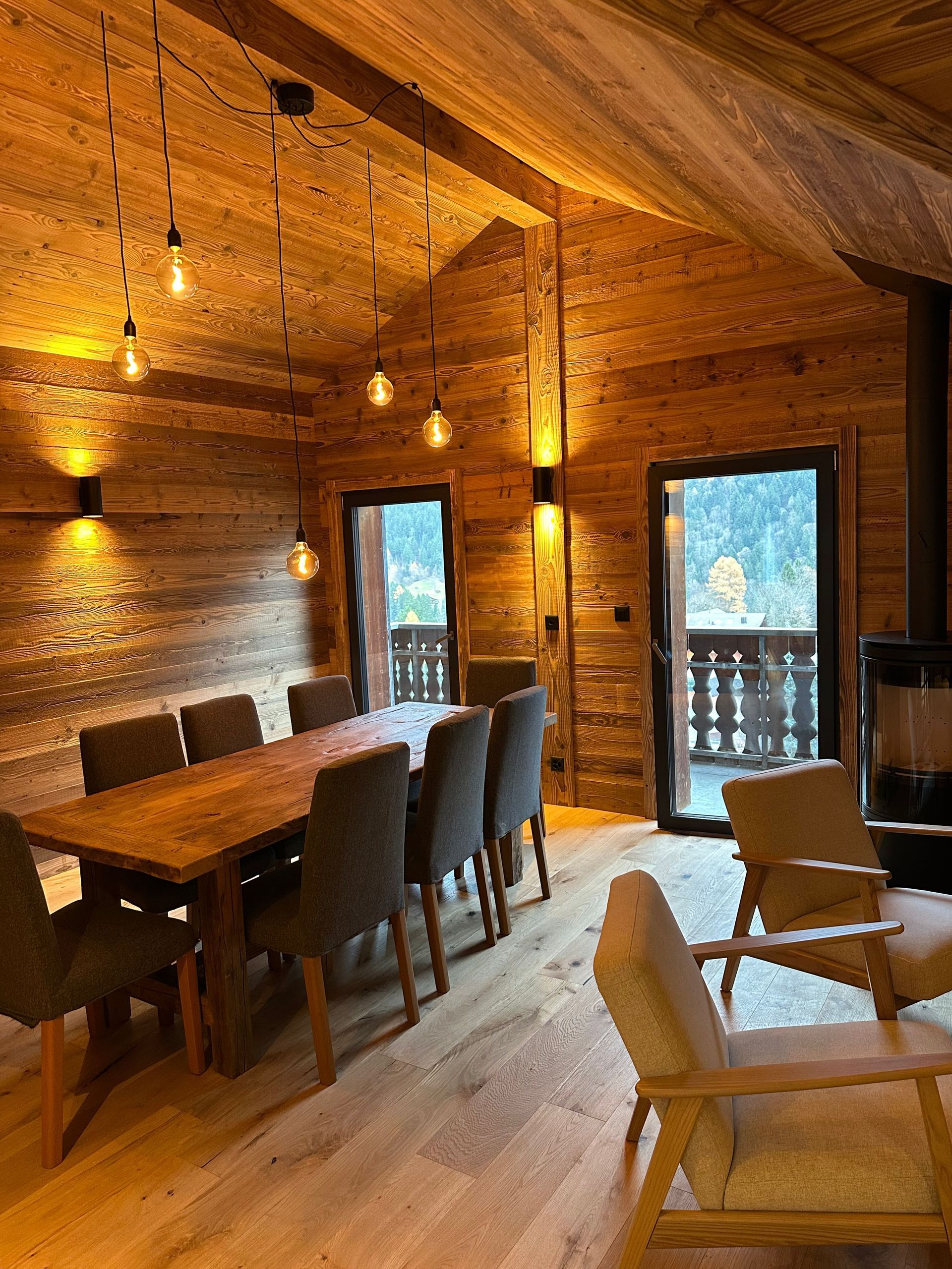 A rustic dining room with wooden walls, a long table, grey chairs, pendant lighting, and two armchairs near a window.