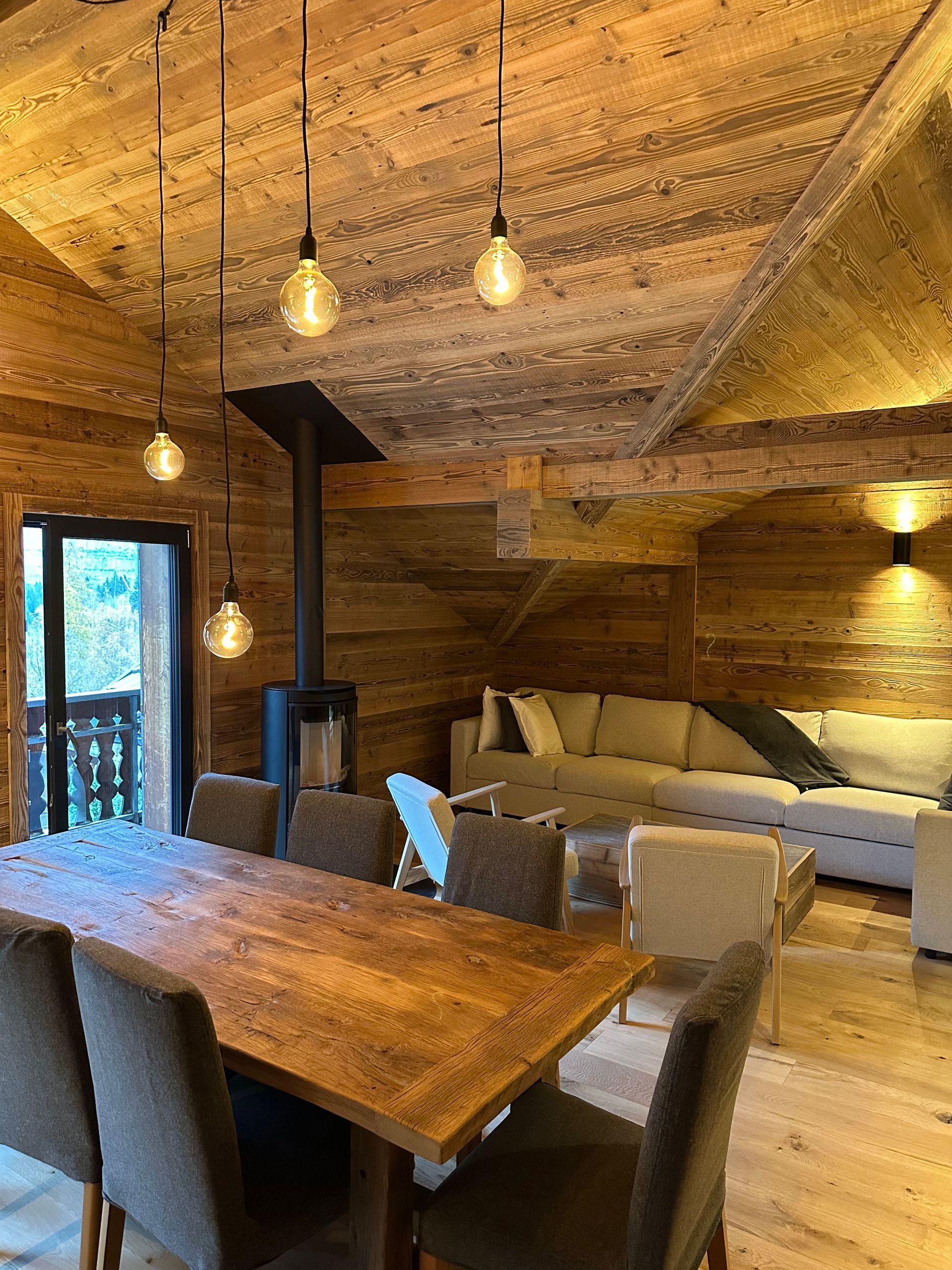 A cozy, rustic room features a wooden dining table, chairs, a wood-burning stove, and a comfortable sectional sofa.