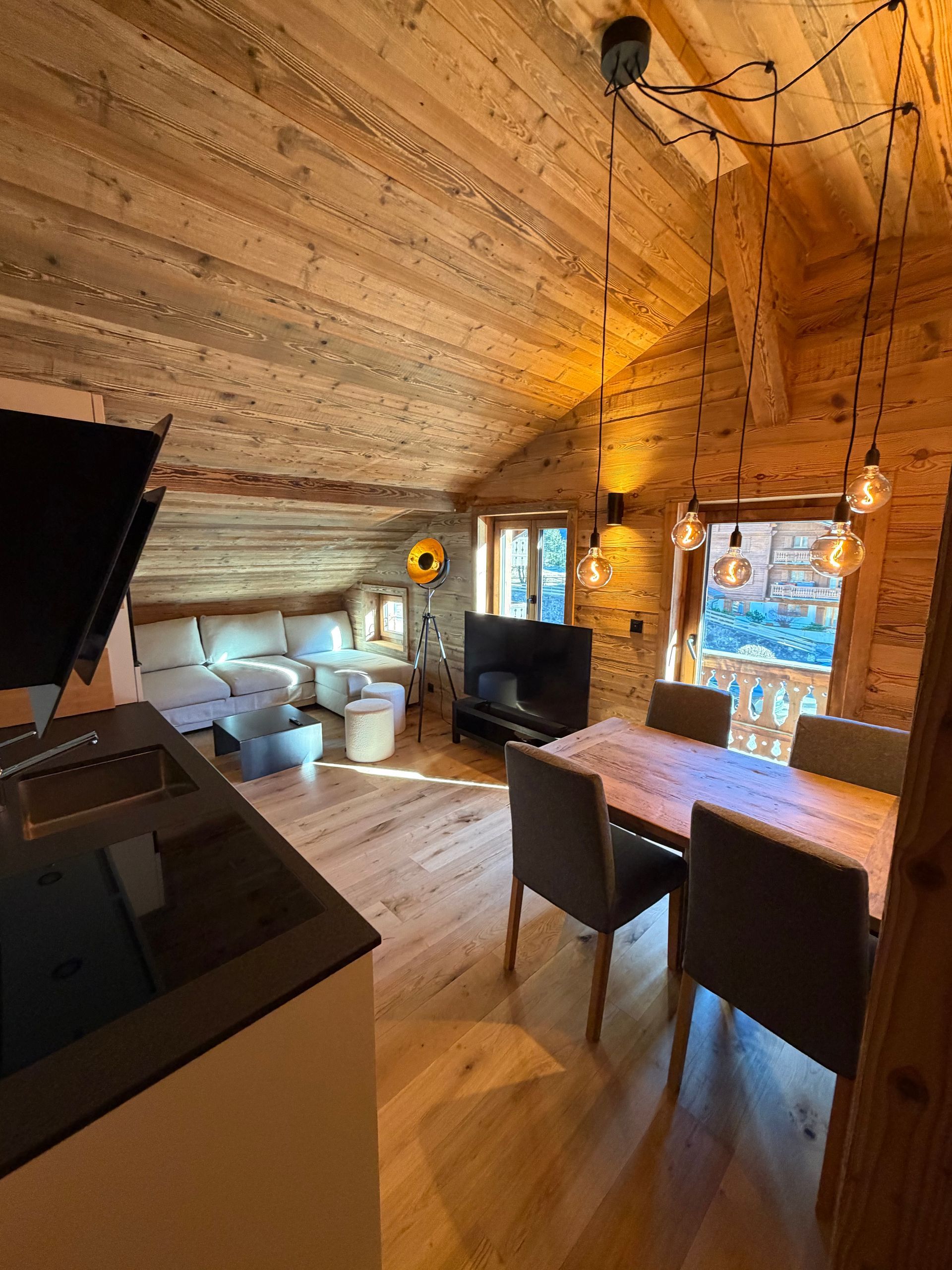 Un intérieur alpin rustique, lambrissé de bois, comprenant une cuisine ouverte, un coin repas avec quatre chaises et un espace de vie confortable.