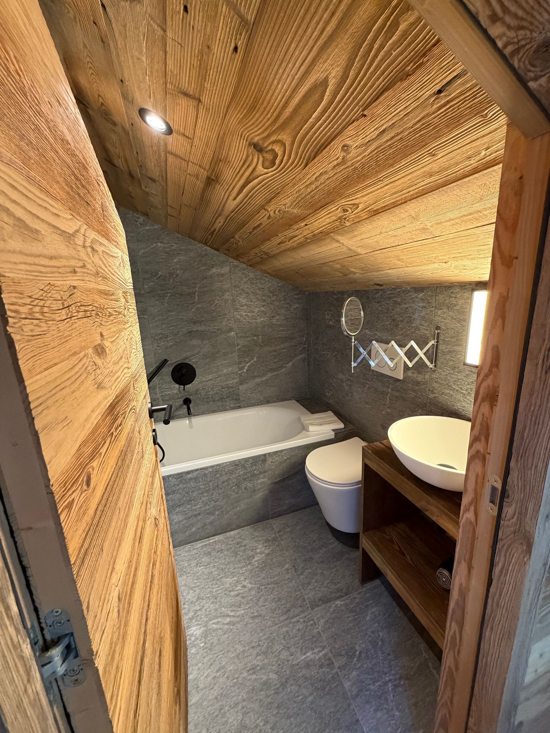 Une petite salle de bains confortable avec un plafond en bois incliné, des murs carrelés gris, une baignoire blanche, des toilettes et un lavabo sur un socle en bois.