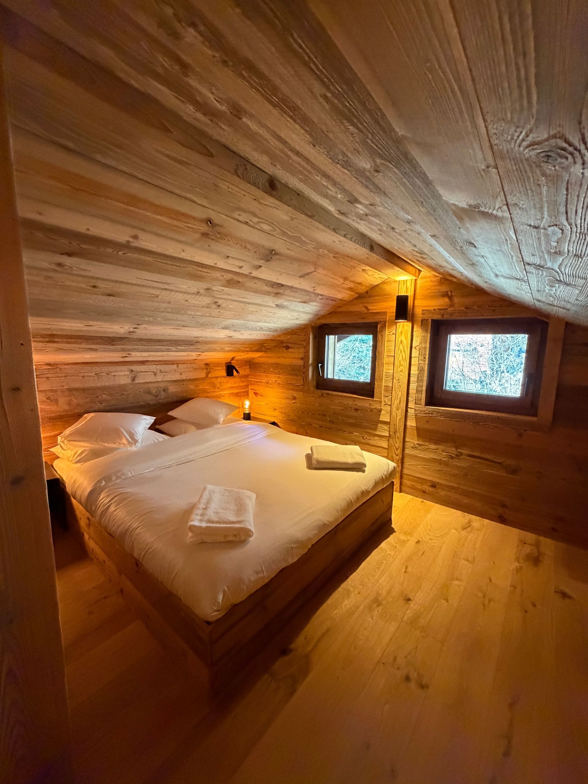 Une chambre confortable avec un plafond et des murs en bois, avec un lit soigneusement fait et des draps blancs sous un toit en pente.