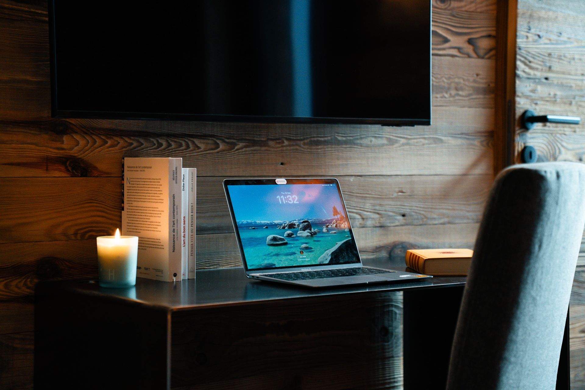 A laptop sits on a dark desk beside a lit candle and books against a rustic wood-paneled wall.