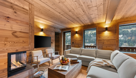 Un salon de chalet moderne, lambrissé, avec une cheminée en pierre, un canapé d'angle beige et des baies vitrées du sol au plafond.