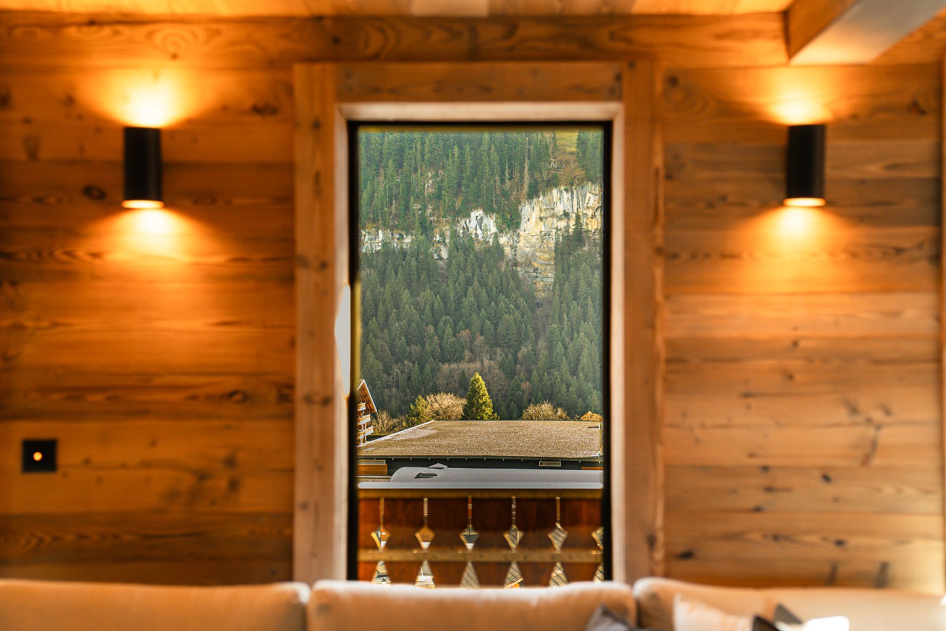 View through a wooden doorway into a balcony overlooking a pine forest and a rocky mountain slope, with two wall sconces.