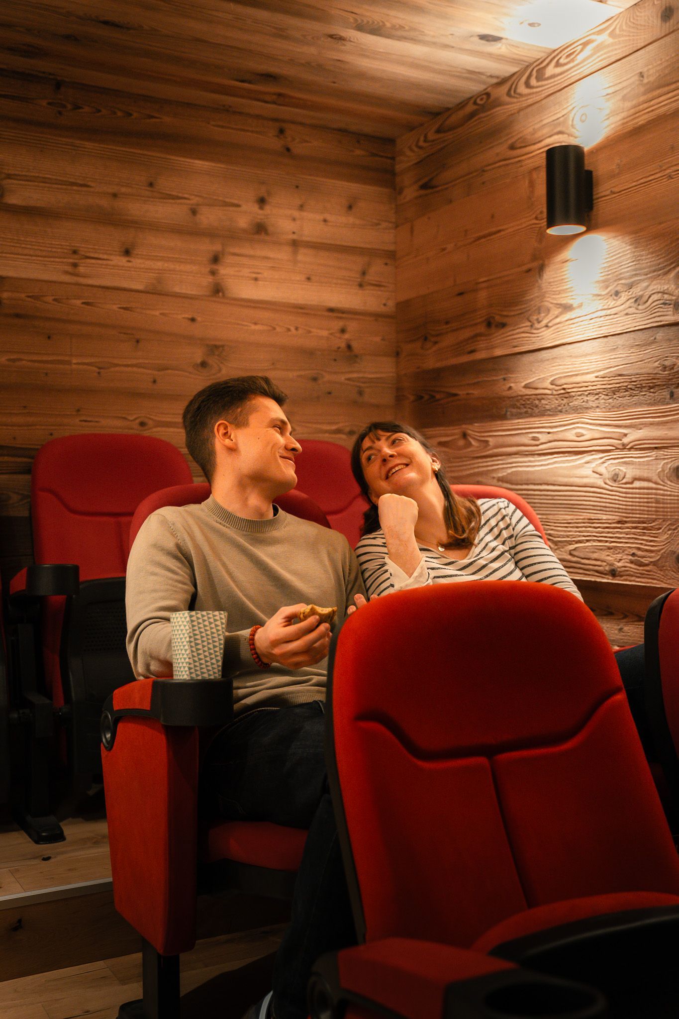 A couple laughs while sitting in red theater chairs in a room with wood-paneled walls and warm lighting.