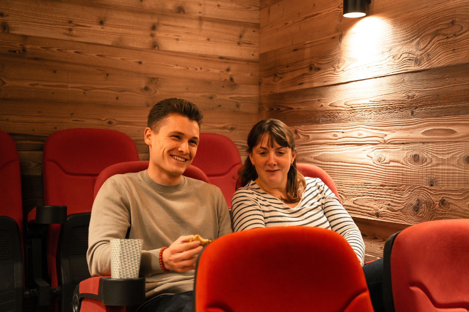 A man and woman sit in red movie theater seats against a wooden wall, smiling while holding snacks.