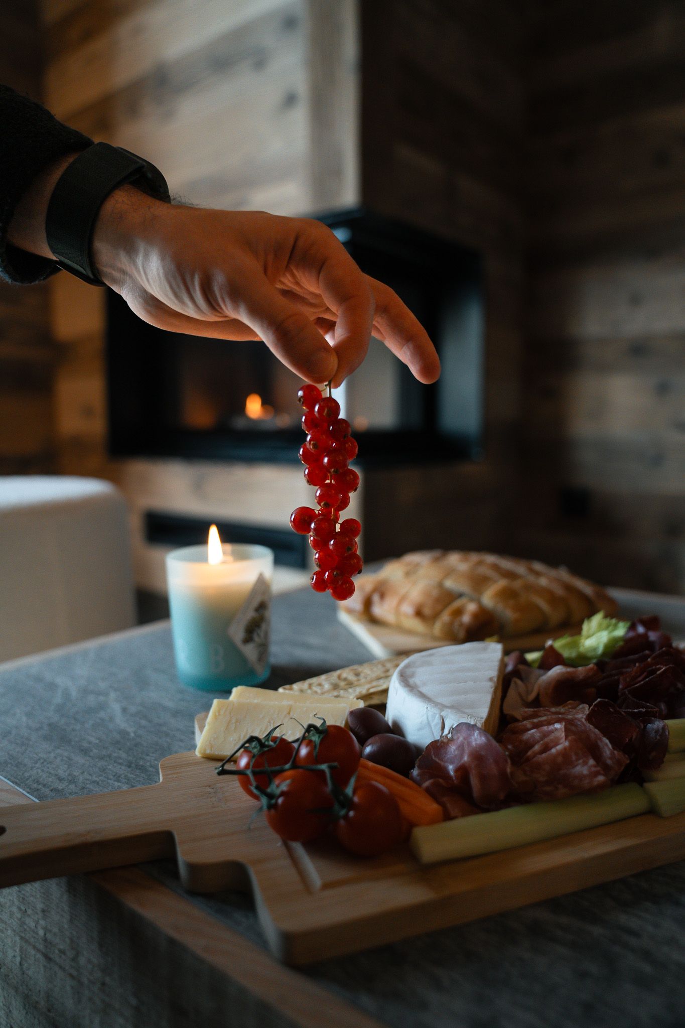 A hand holds a stem of red currants above a rustic wooden charcuterie board featuring cheese, bread, and meats near a candle.