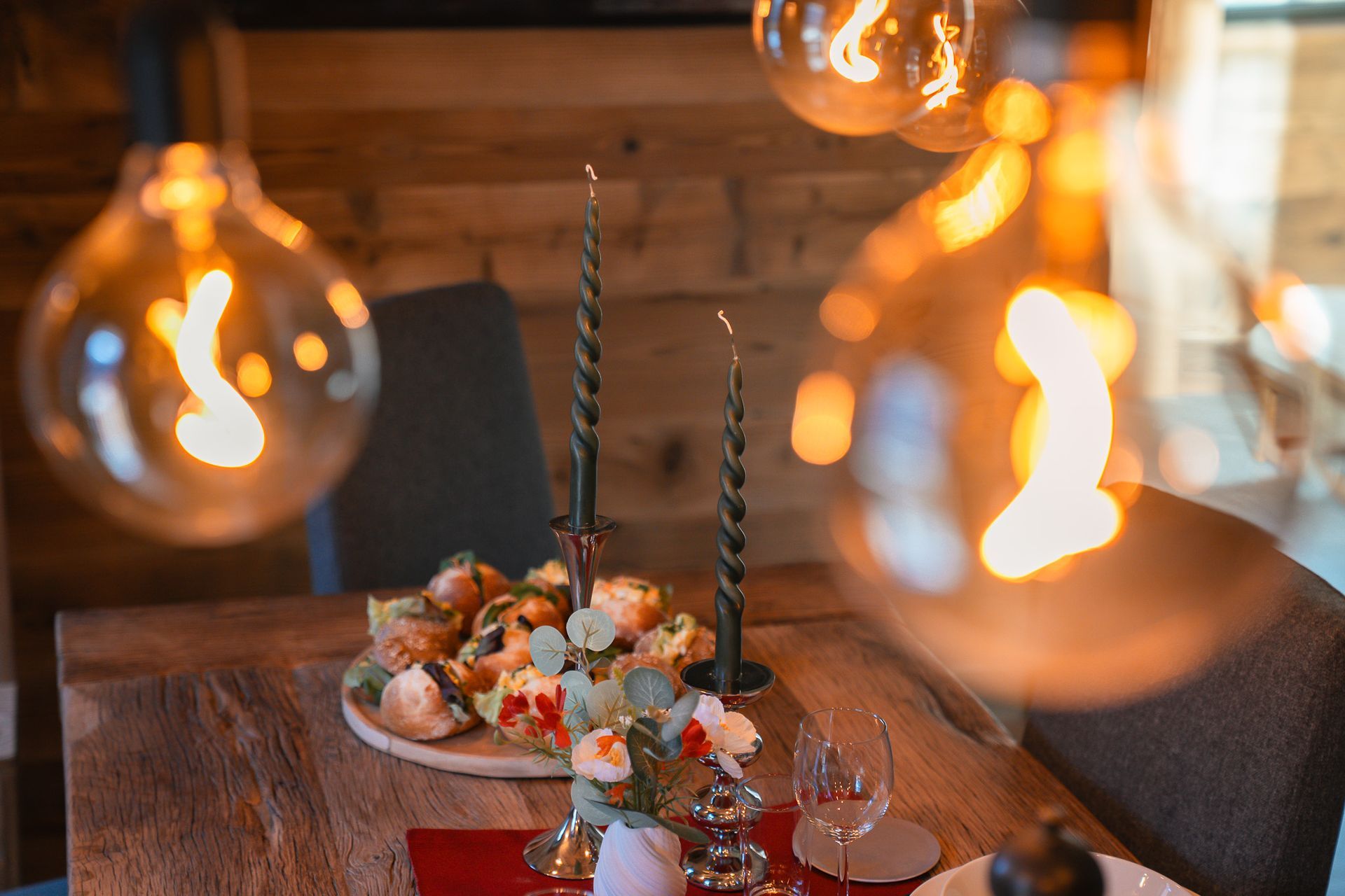 A rustic wooden dining table features a decorative centerpiece with candles and foliage, illuminated by warm globe lights.