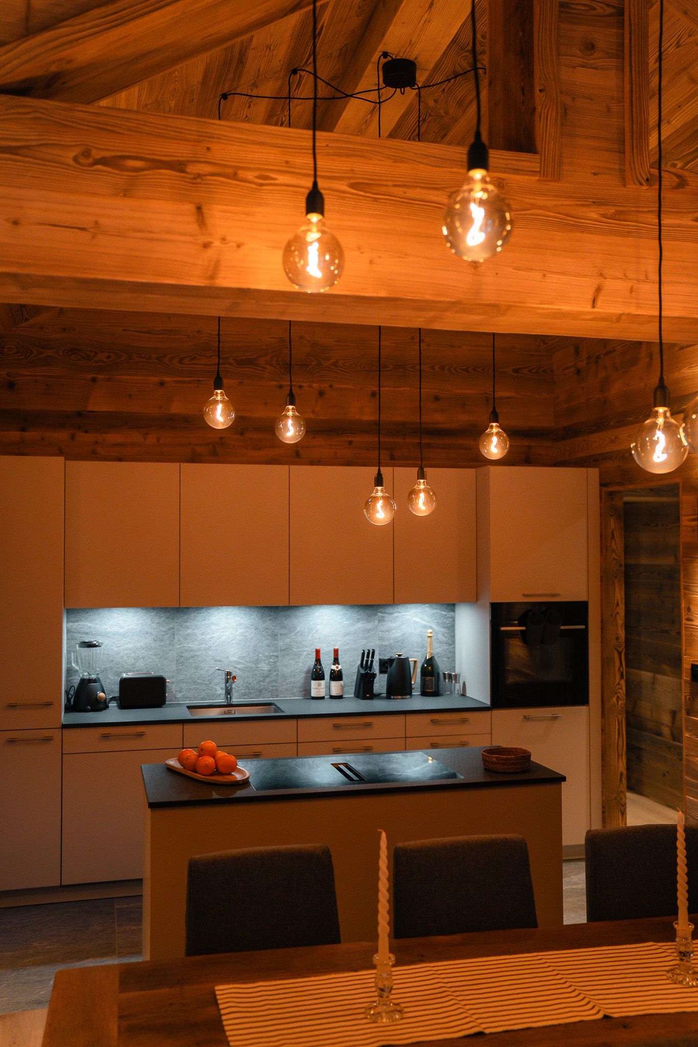 A rustic kitchen with wood beams and modern white cabinetry, lit by warm hanging filament bulbs above an island.
