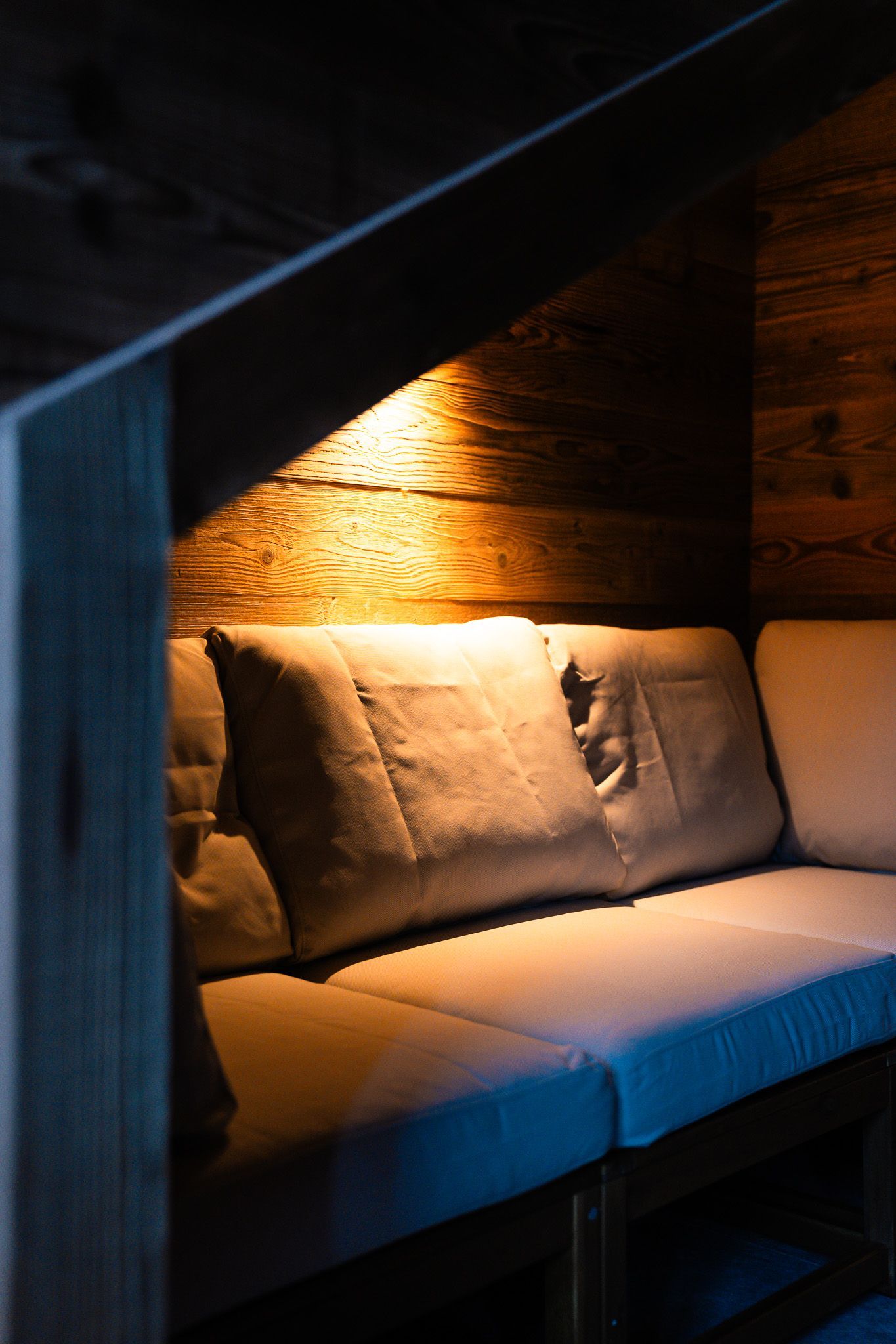 Cozy seating area with light-colored cushions against a rustic wooden wall, illuminated by warm, soft lighting.