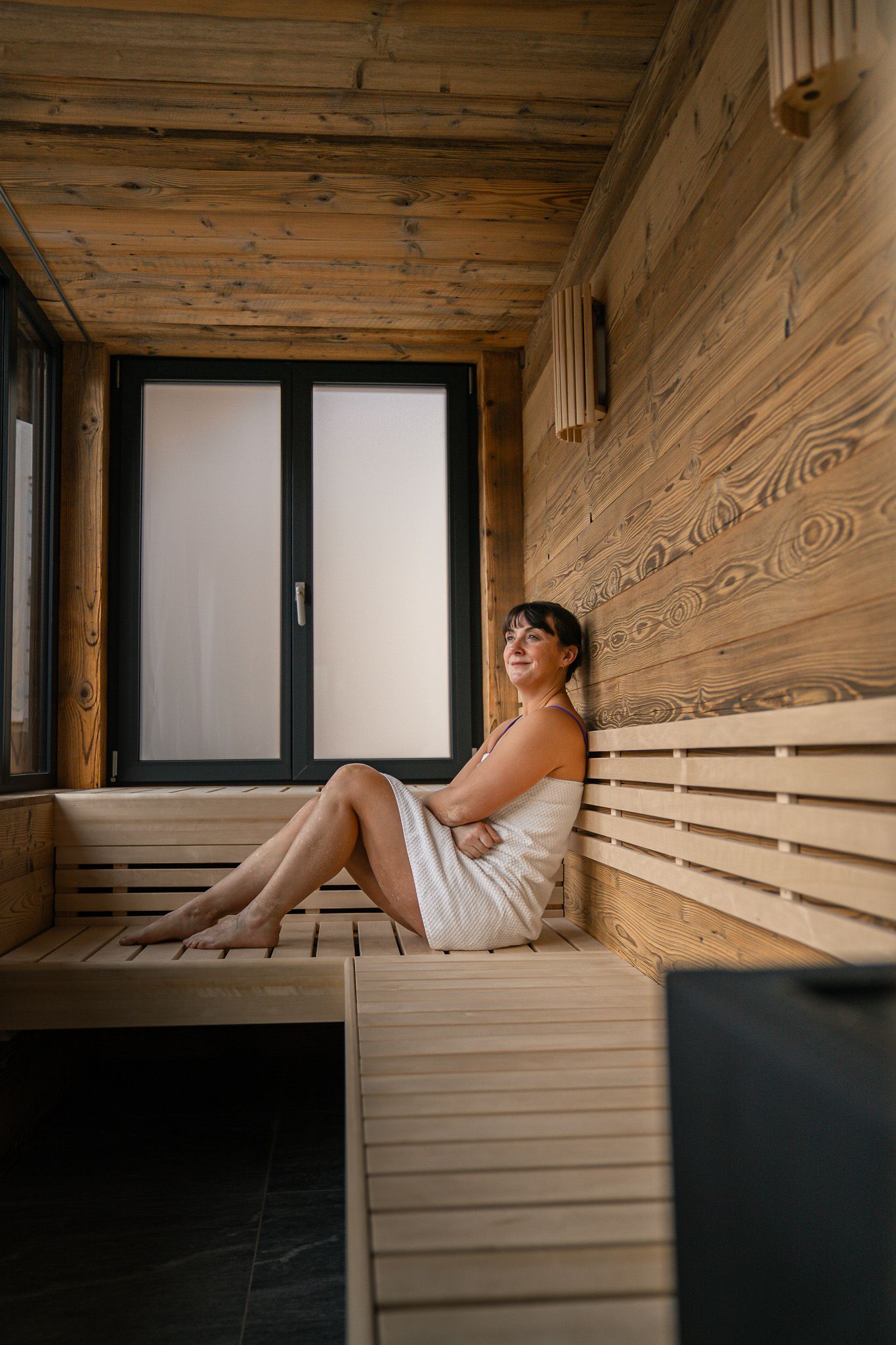 A woman in a white towel sits relaxing in a wooden sauna with a frosted window.