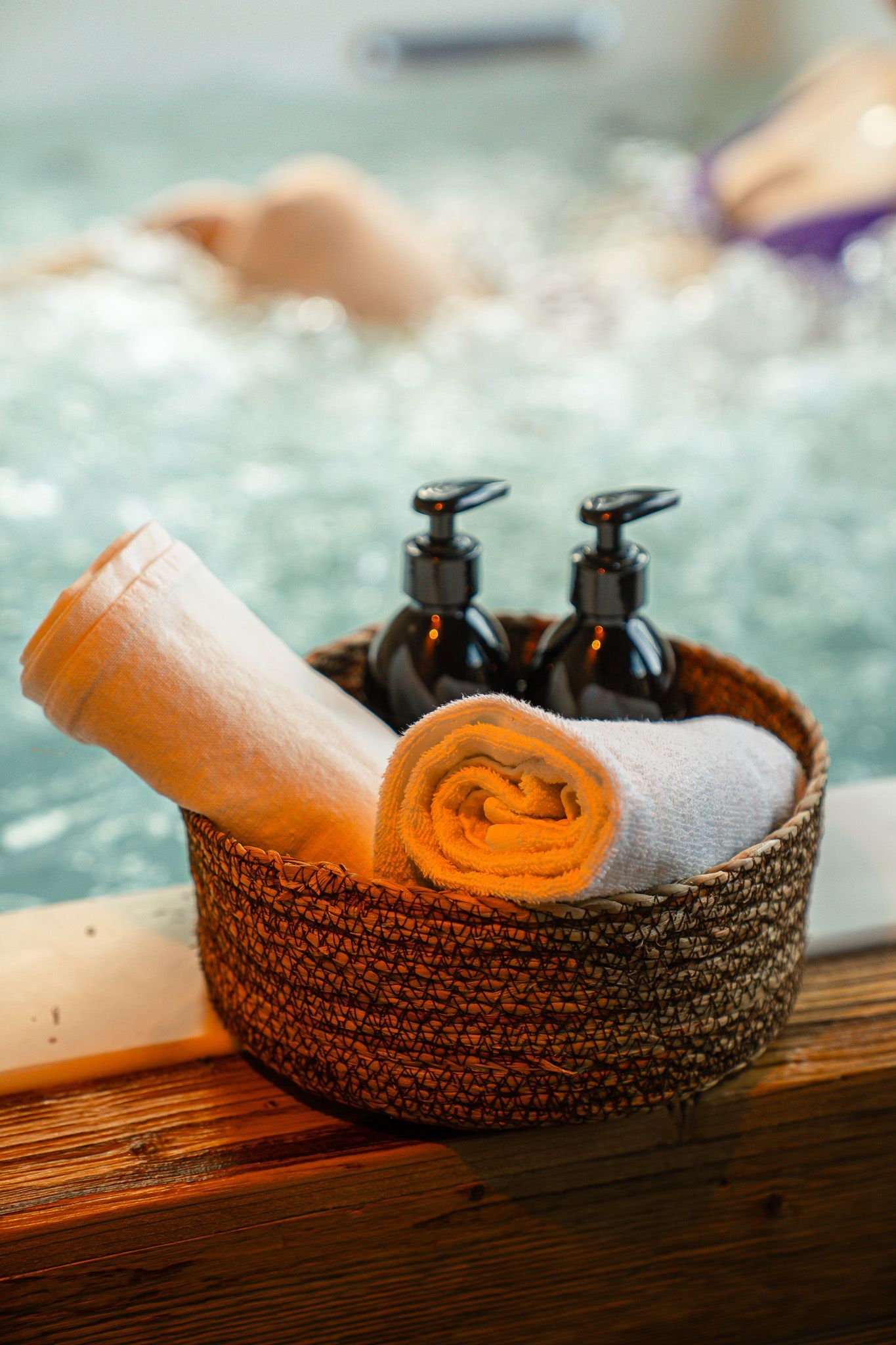 A woven basket containing two rolled towels and two pump bottles sits on a wooden ledge beside a bubbling hot tub.