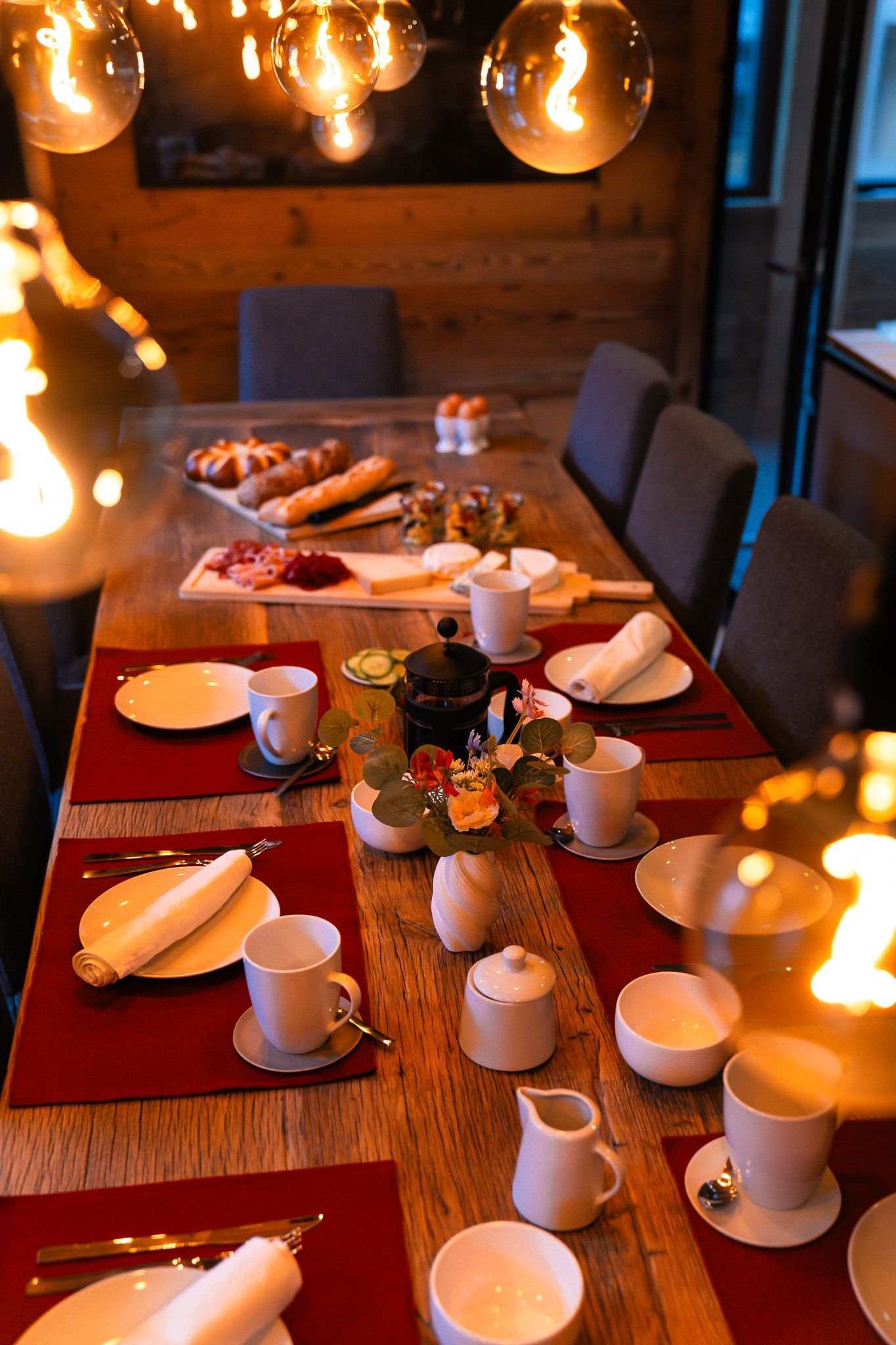 A rustic wooden dining table set for breakfast with white dishes, red placemats, and a platter of food, lit by warm bulbs.