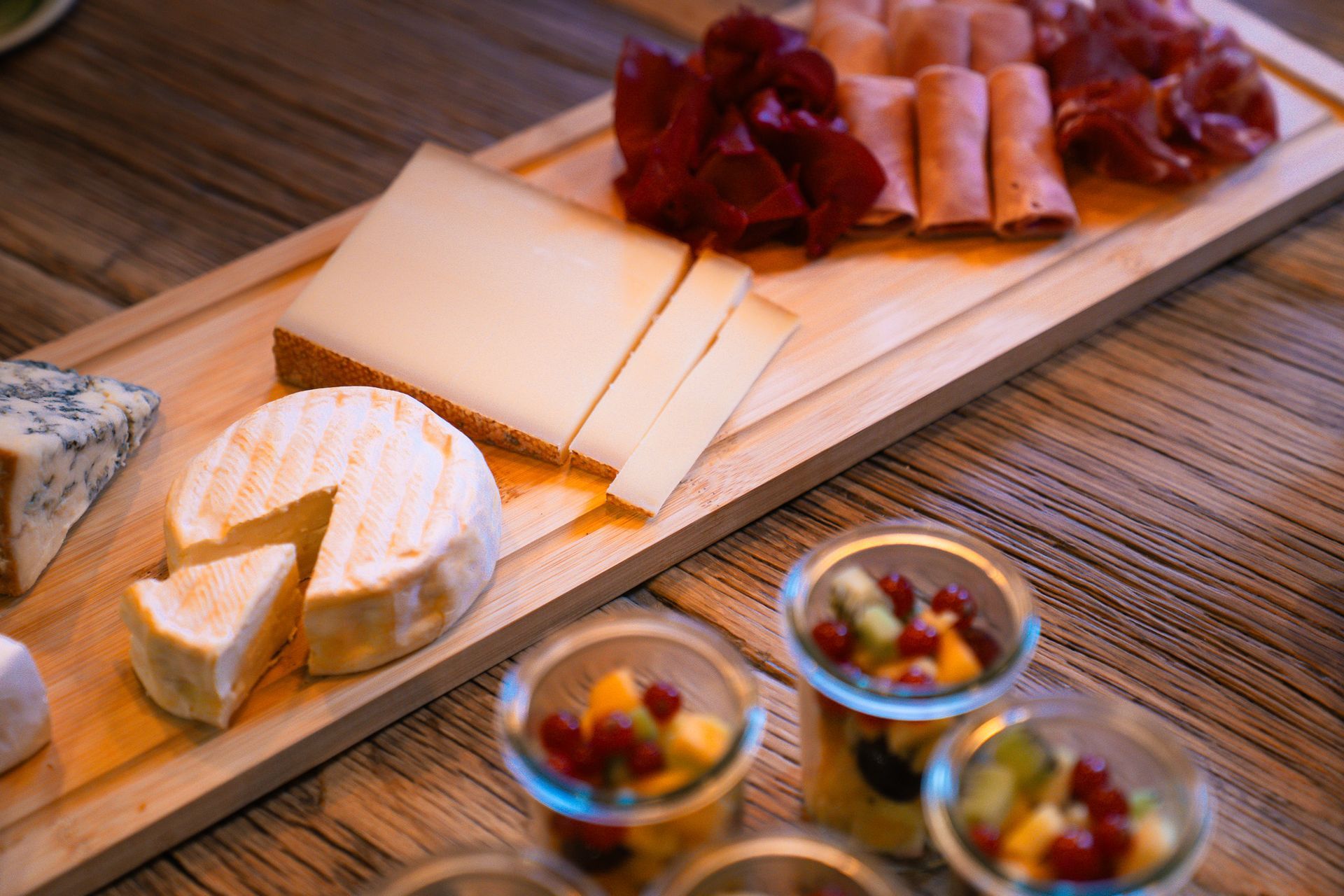 A wooden board displays various cheeses, including blue cheese and a sliced round, next to folded deli meats and fruit cups.