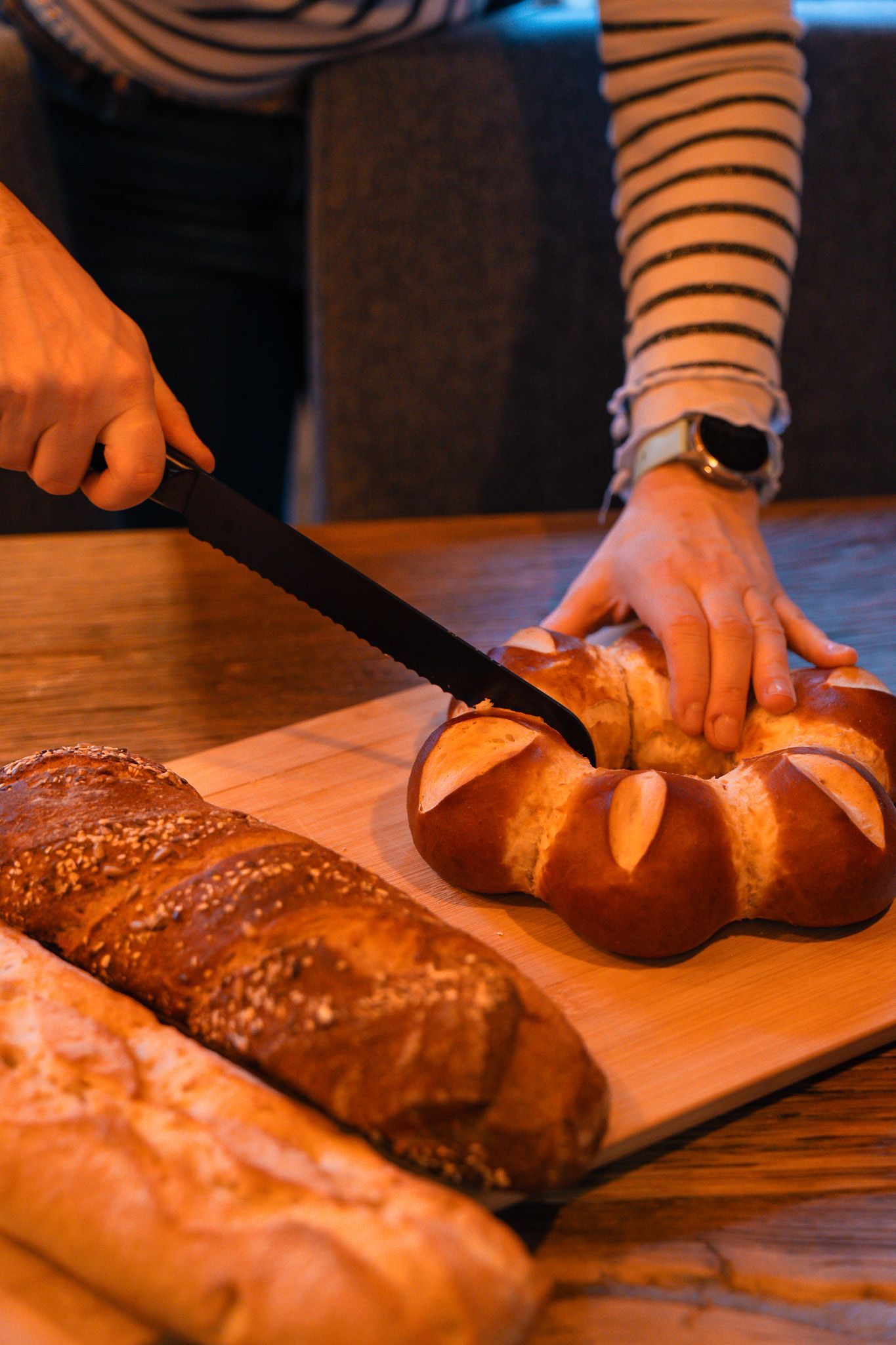 A person in a striped shirt cuts a circular pretzel-style loaf of bread on a wooden board next to a baguette.