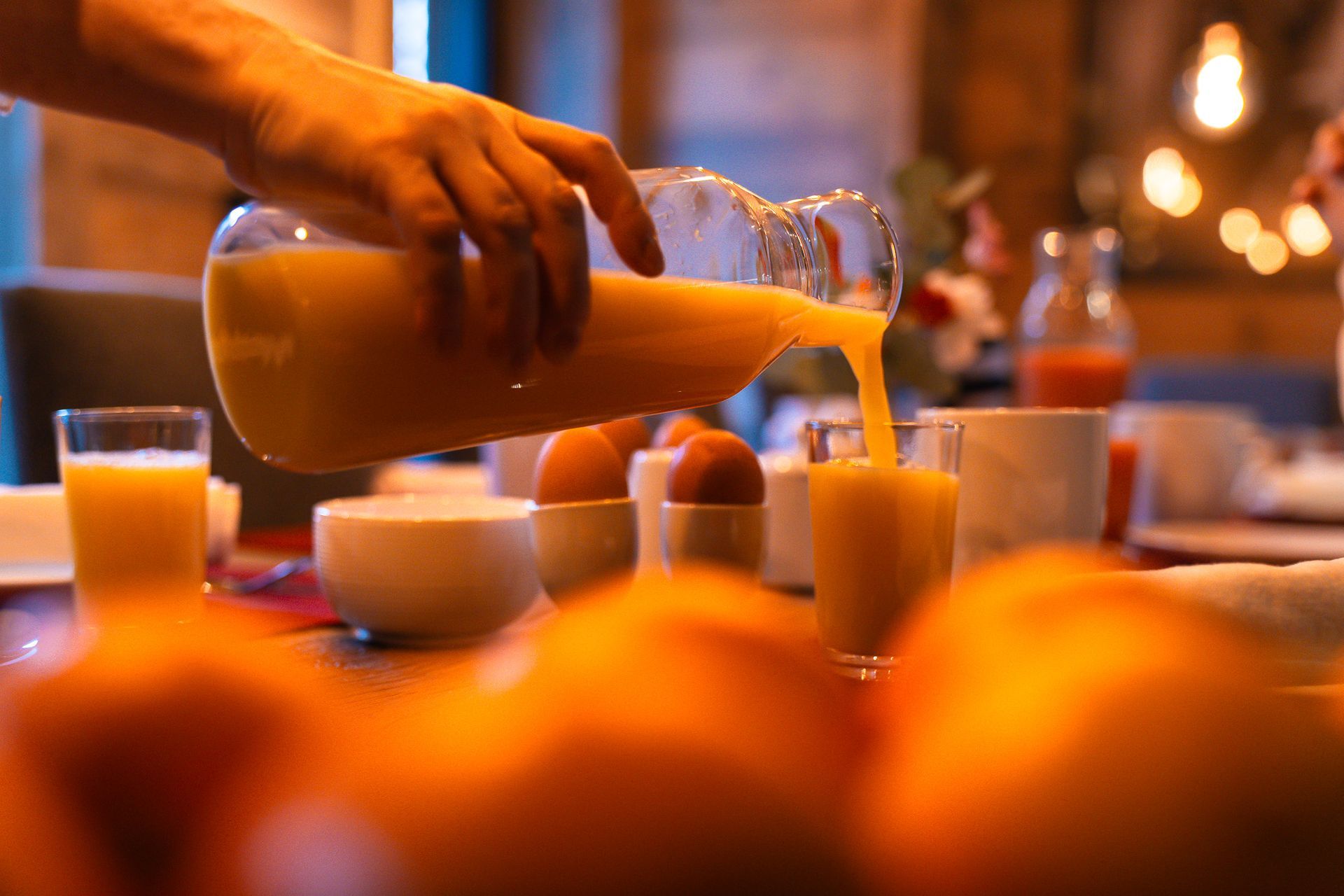 Une main verse du jus d'orange d'un pichet en verre dans un verre posé sur une table de petit-déjeuner où se trouvent également des œufs et des bols.