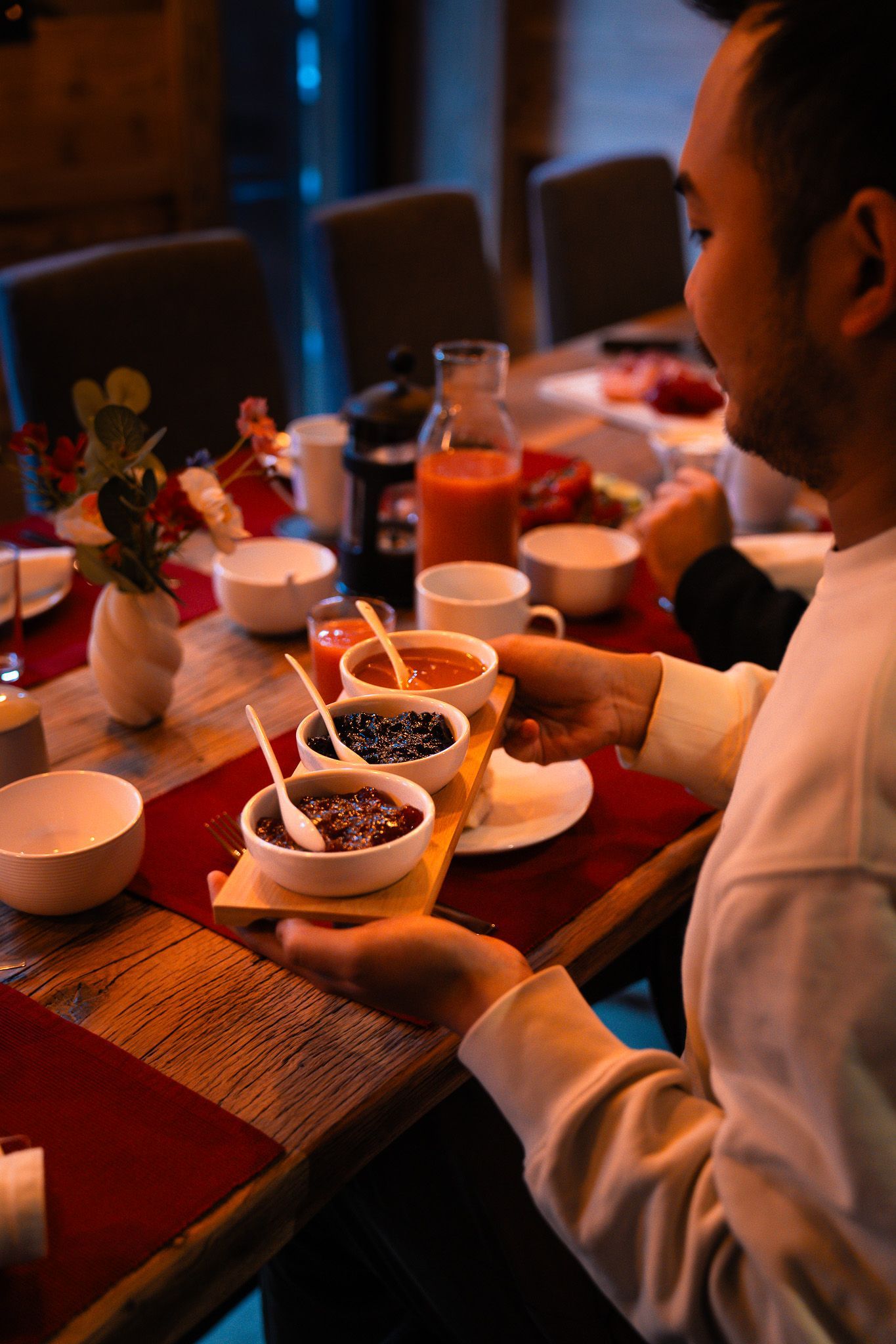 Dans une pièce faiblement éclairée, une personne tient une planche en bois sur laquelle sont disposés trois petits bols de nourriture colorée, autour d'une table dressée pour un repas.