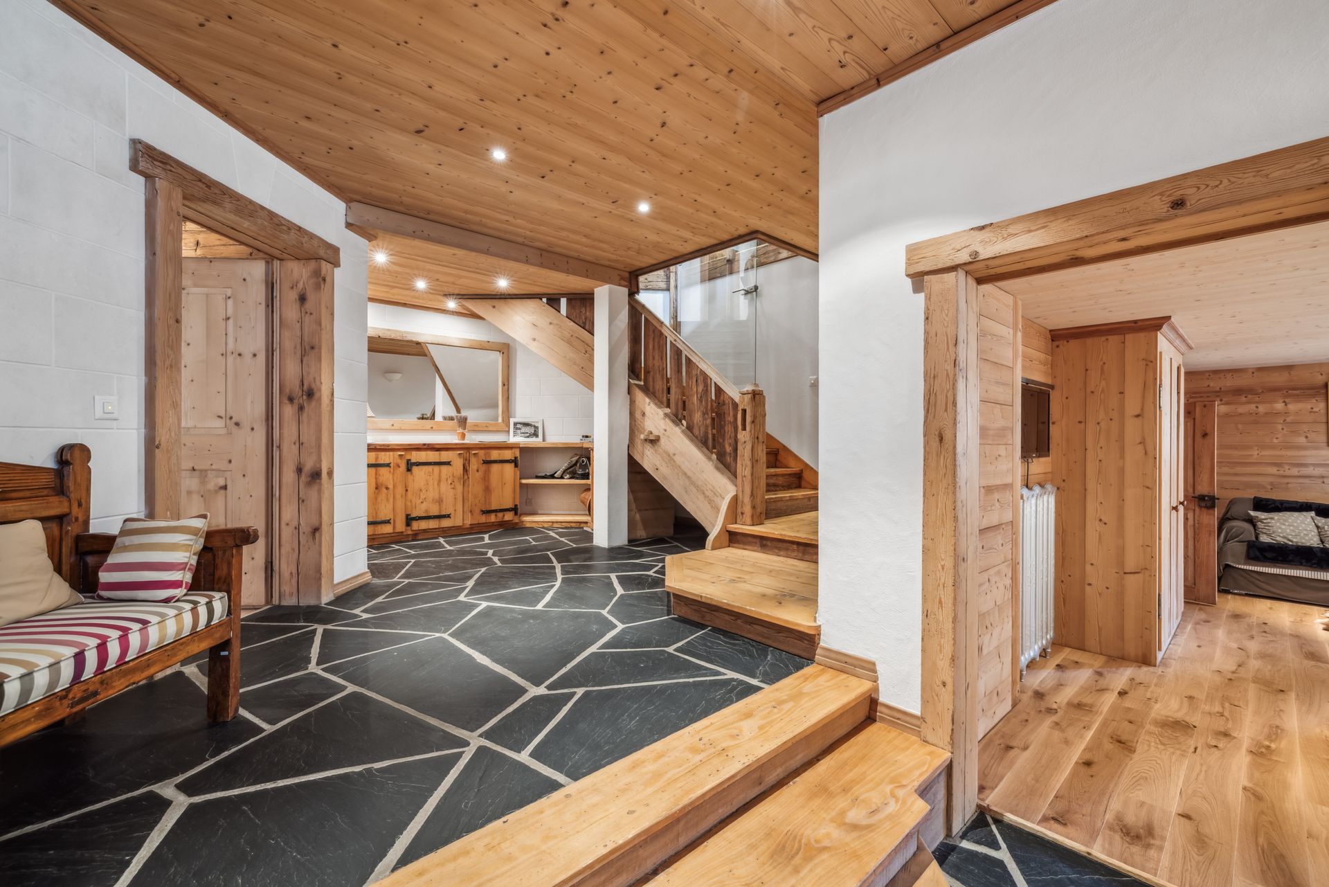 A rustic entryway featuring a dark stone floor, wooden stairs, a bench, and wood-paneled walls and ceilings.