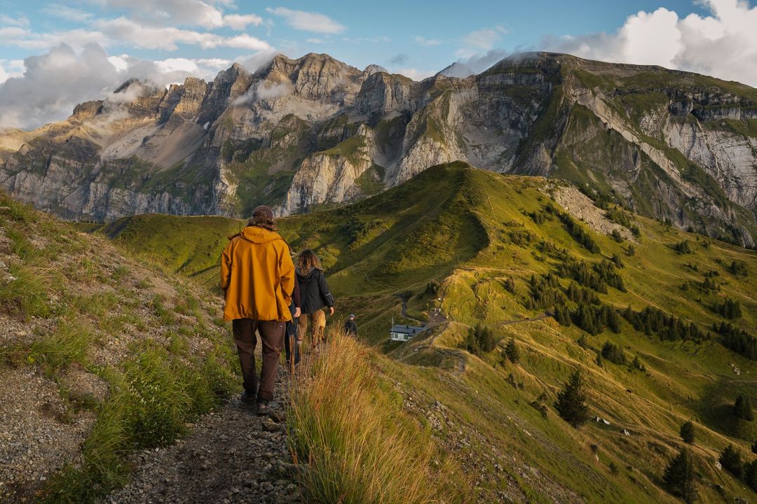 Deux personnes randonnent sur un sentier de montagne étroit et herbeux, en direction de pics rocheux, sous un ciel partiellement nuageux.