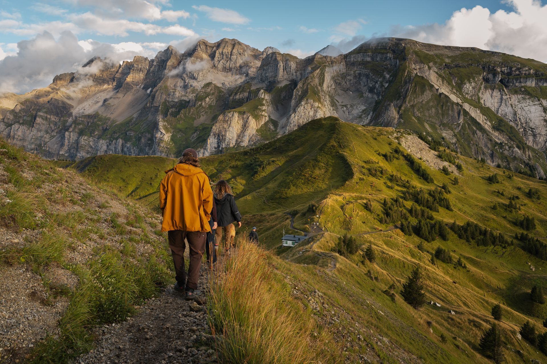 Two people hike along a rugged mountain trail with lush green slopes and dramatic, rocky peaks in the background.