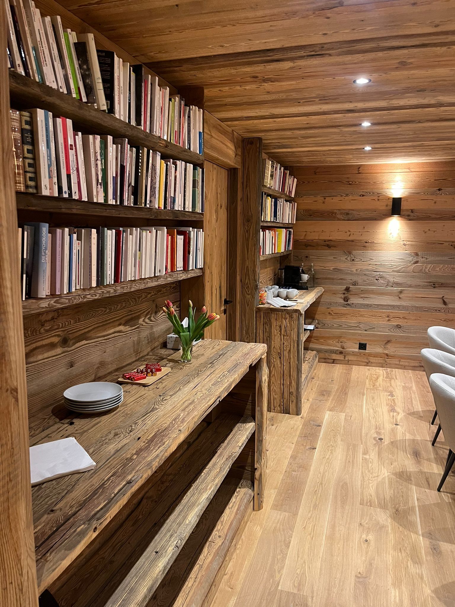 Rustic library with wooden shelving, walls, and ceiling, featuring many books and a long wooden counter in a warm room.