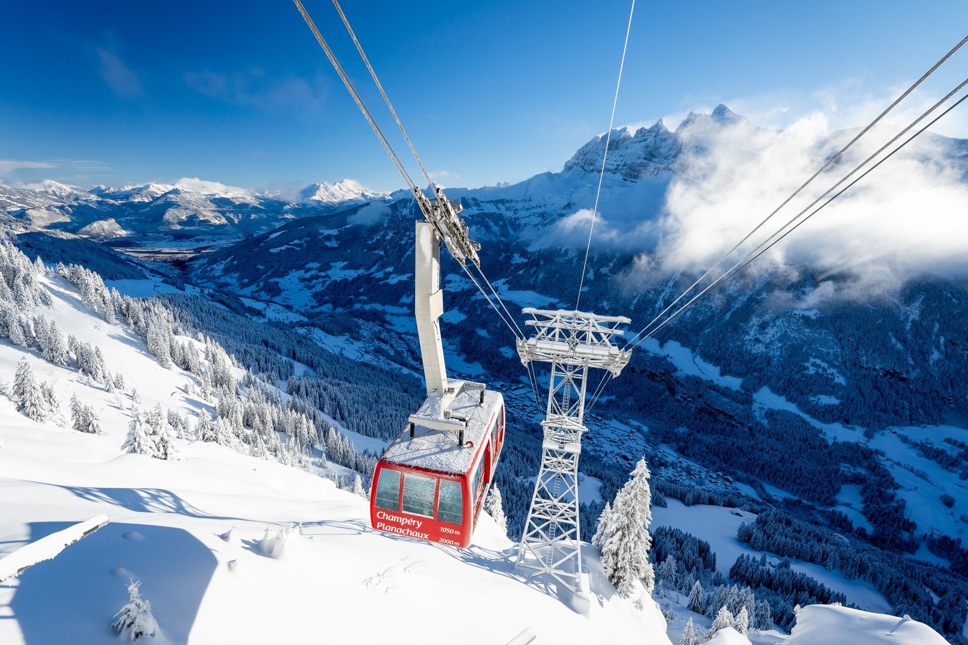 A bright red cable car travels on cables above a snowy mountain landscape under a clear blue sky.