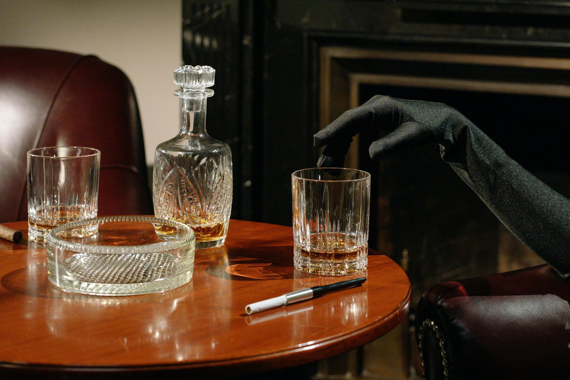 Une main gantée tend la main vers un verre de whisky posé sur une table avec une carafe, un cendrier et un cigare.