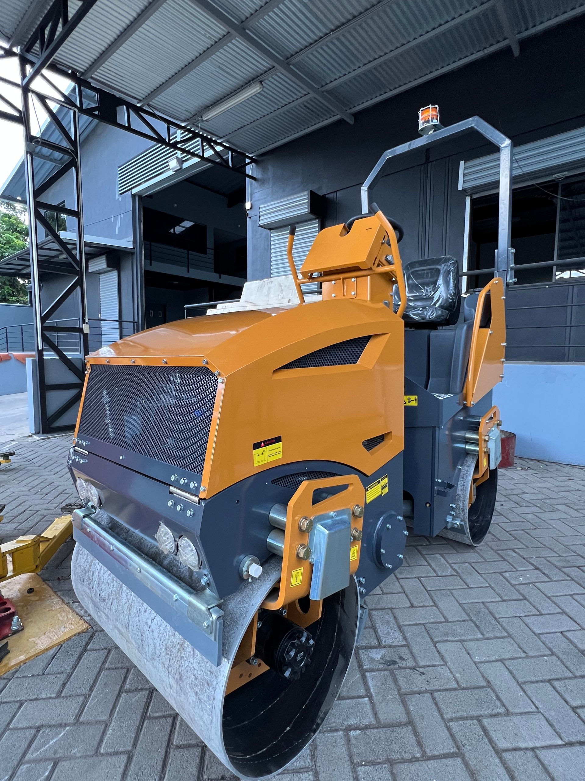 Un rodillo compactador sobre un suelo de ladrillo, con un cuerpo de color naranja y gris, debajo de un área cubierta.