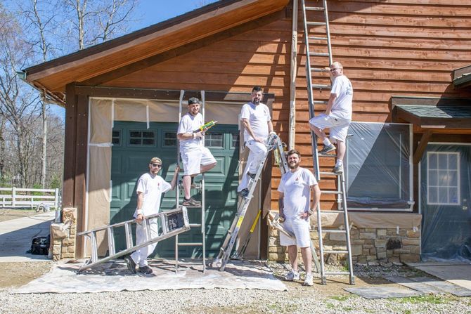 Five workers painting a wooden building exterior, with ladders and tools outside in daylight.