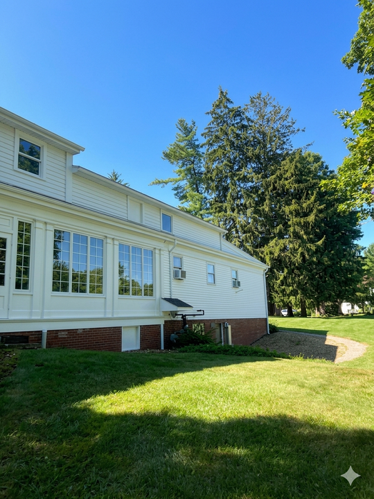 White house exterior with large windows, brick foundation, and a sunny green lawn beside tall trees
