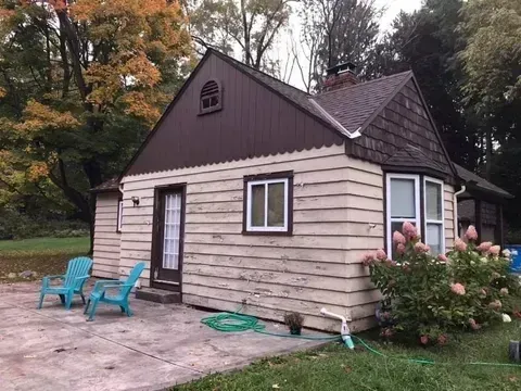 Small beige cabin with dark roof, turquoise chairs on the patio, and flowers by the entrance